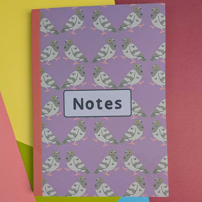 A purple notebook with a repeating pattern of cartoon pigeons and a white label reading 'Notes' on the front, photographed on a colorful background of pink, yellow, and green paper.