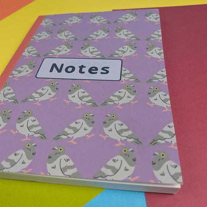 A purple notebook with a repeating pattern of gray and white pigeons and a white label reading 'Notes' on the front, photographed on a colorful background.