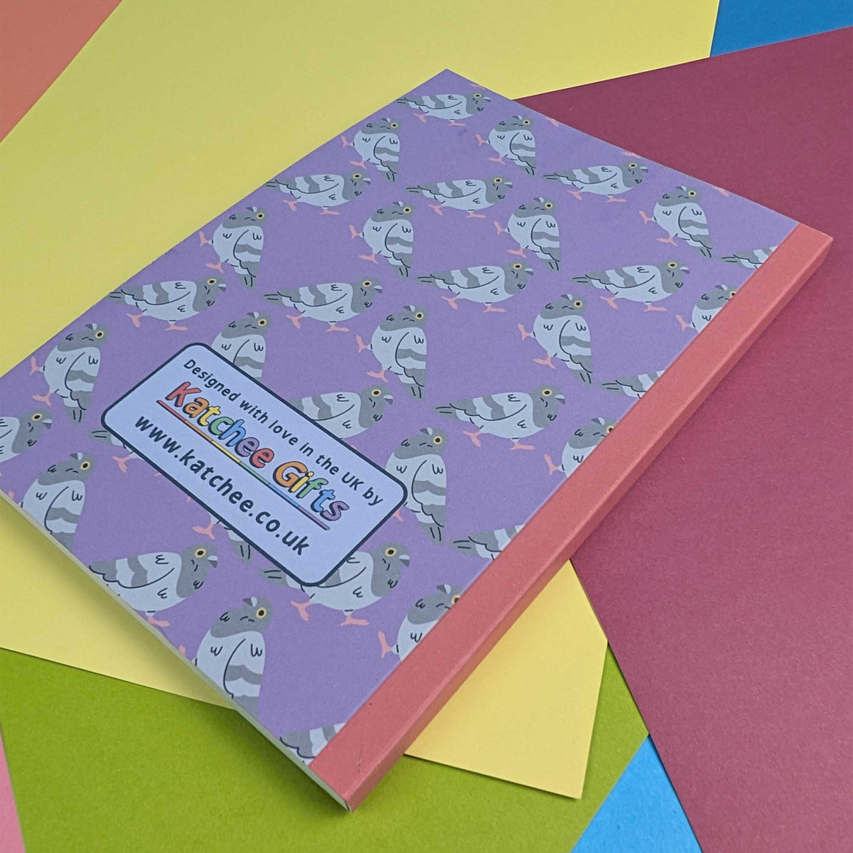 A purple notebook or journal with a repeating pattern of gray pigeons, placed on top of colorful paper sheets. A white label on the cover reads 'Designed with love in the UK by Katchee Gifts' 