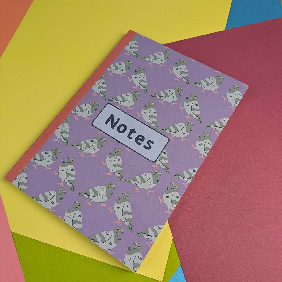 A purple 'Notes' notebook with a repeating pattern of cartoon pigeons, styled on top of a stack of colorful paper sheets.