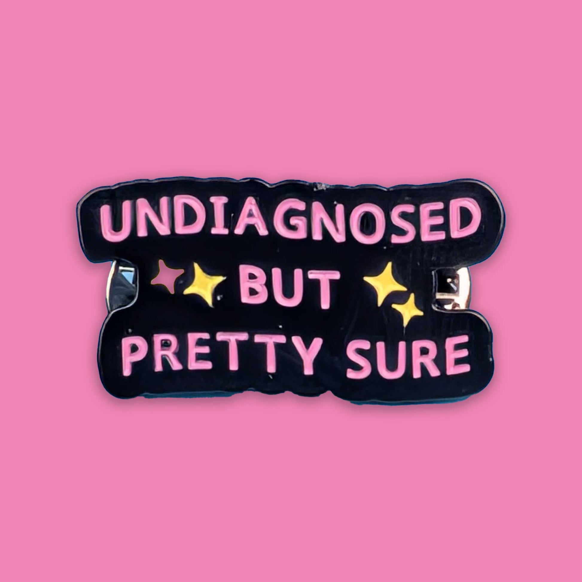 A black enamel pin with the text 'UNDIAGNOSED BUT PRETTY SURE' in pink letters, accented with yellow stars, shown on a solid pink background.