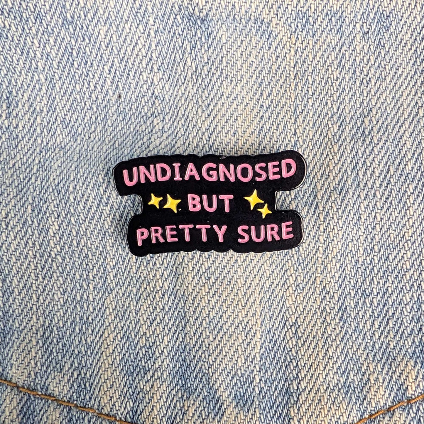 A small enamel pin with the text 'UNDIAGNOSED BUT PRETTY SURE' in pink letters on a black background with yellow stars, placed on a light blue denim fabric.