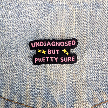 A small enamel pin with the text 'UNDIAGNOSED BUT PRETTY SURE' in pink letters on a black background with yellow stars, placed on a light blue denim fabric.