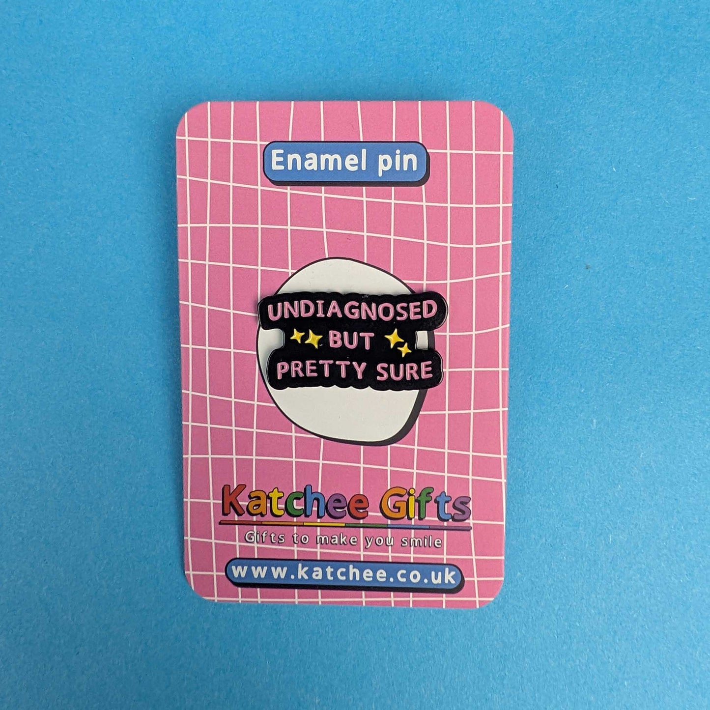 A pink enamel pin on its retail backing card, photographed against a solid blue background. The backing card features a pink grid pattern, a blue label reading 'Enamel pin' at the top, and the brand name 'Katchee Gifts' at the bottom.