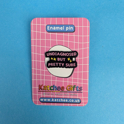 A pink enamel pin on its retail backing card, photographed against a solid blue background. The backing card features a pink grid pattern, a blue label reading 'Enamel pin' at the top, and the brand name 'Katchee Gifts' at the bottom.