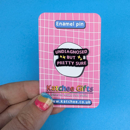A hand holds a pink enamel pin backing card featuring a white grid pattern and the text 'Enamel pin' at the top. The backing card displays a black enamel pin with a white oval in the center that reads 'UNDIAGNOSED BUT PRETTY SURE' in black and yellow lettering.