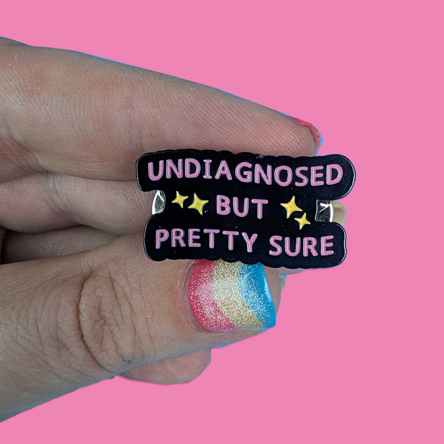 A hand with glittery rainbow nail polish holds a black enamel pin that reads 'UNDIAGNOSED BUT PRETTY SURE' in pink letters with yellow stars, set against a solid pink background.