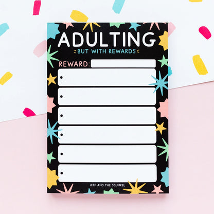 Notepad with 'Adulting But with Rewards' text and colorful stars on a light pink background