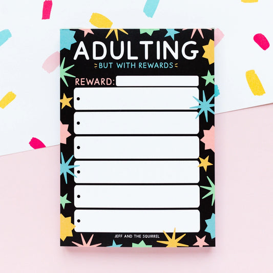 Notepad with 'Adulting But with Rewards' text and colorful stars on a light pink background