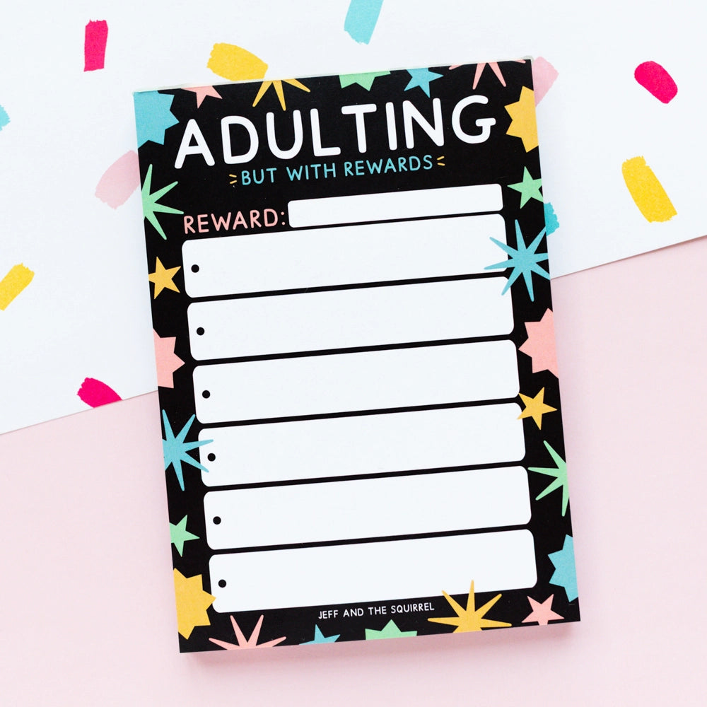 Notepad with 'Adulting But with Rewards' text and colorful star patterns on a light pink background.