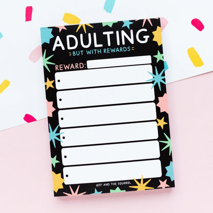 Notepad with 'Adulting But with Rewards' text and colorful star patterns on a light pink background.