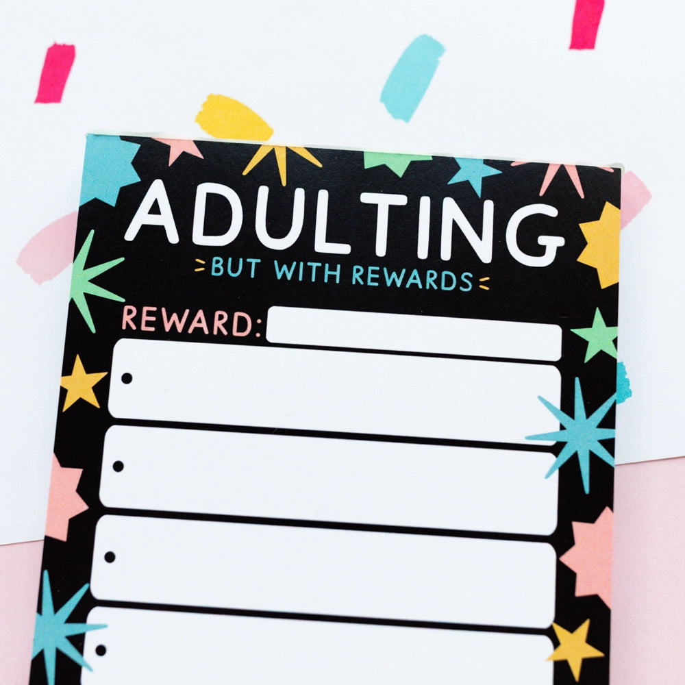 A rectangular reward card with a black background and colorful starburst and star patterns around the border. Large white text reads 'ADULTING' with smaller teal text 'BUT WITH REWARDS' above a blank 'REWARD:' field and several lined boxes underneath.