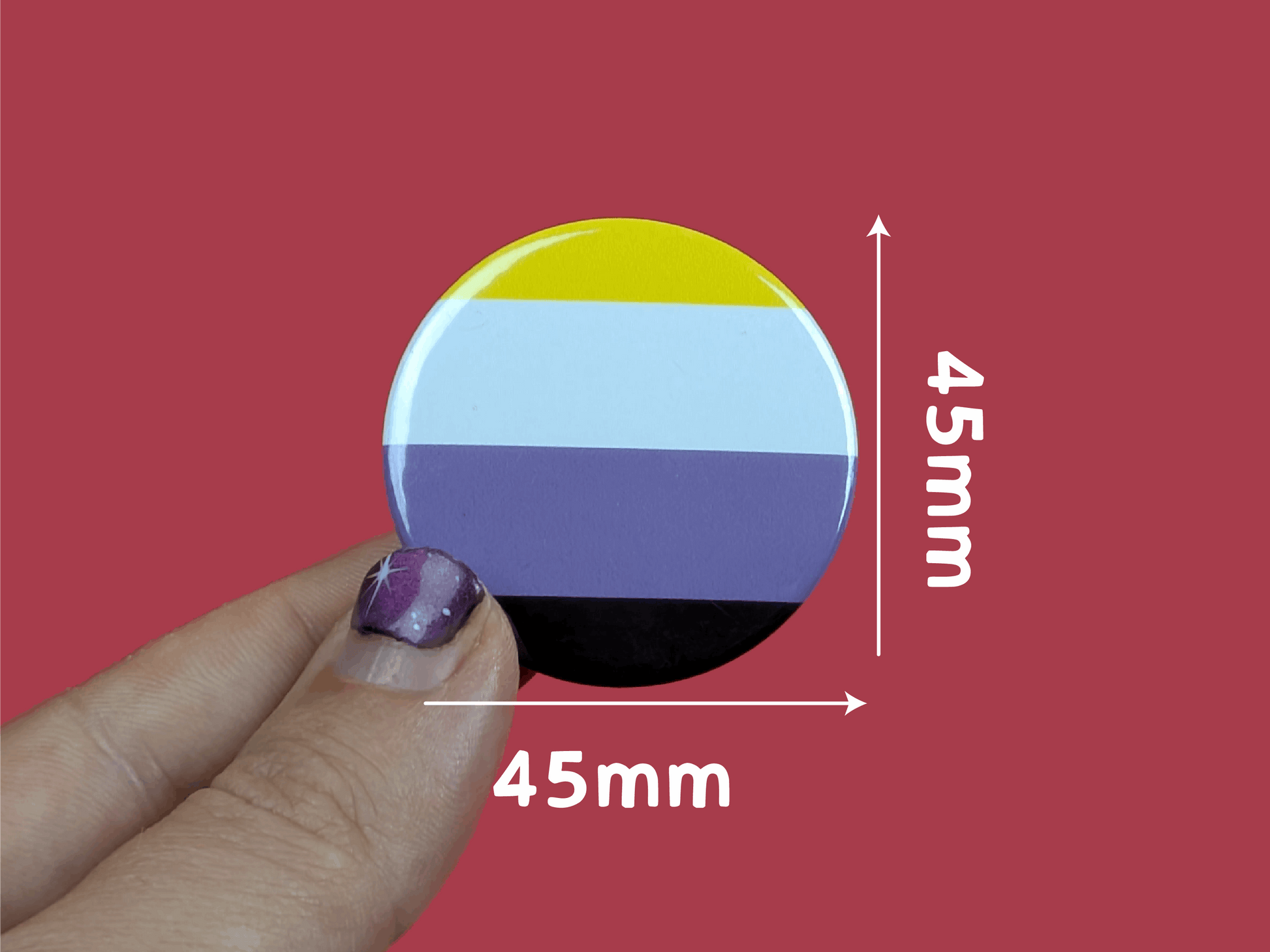Round badge with a non binary flag design held by a hand against a red background, with measurements indicated.