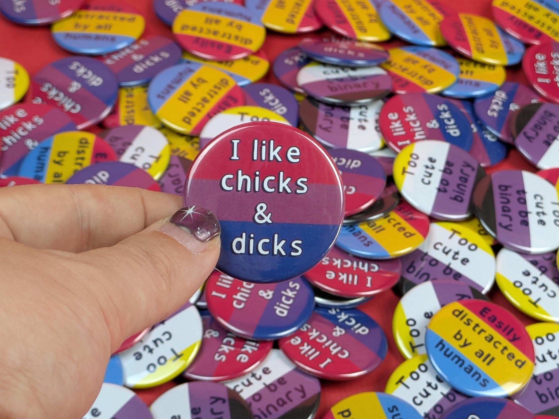 Collection of buttons with various text, with a hand holding one that says 'I like chicks & dicks'.