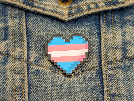 Pixelated heart-shaped pin with a transgender flag design on a denim jacket.