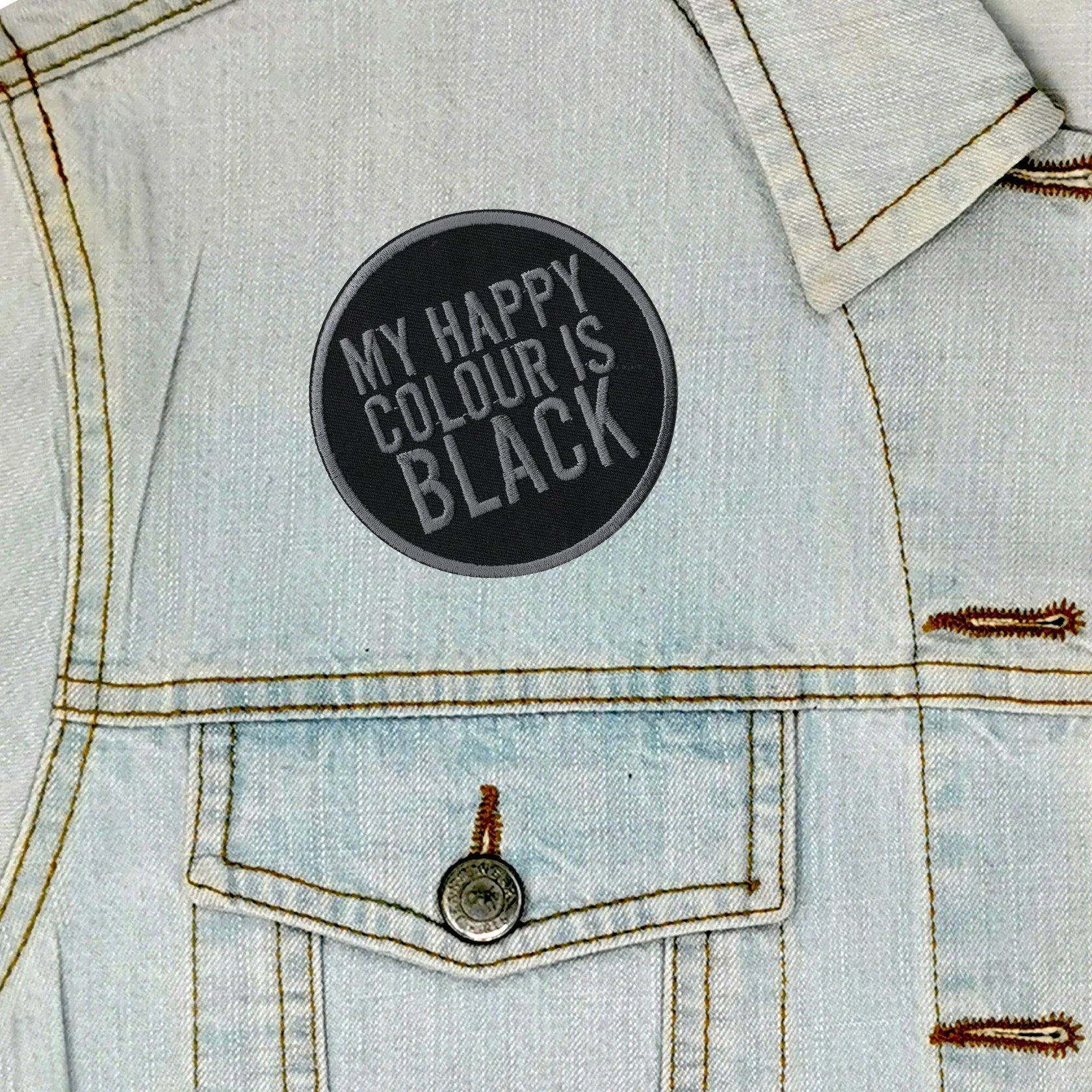 A circular black patch with the text 'MY HAPPY COLOUR IS BLACK' in gray lettering, shown on the chest of a light blue denim jacket.