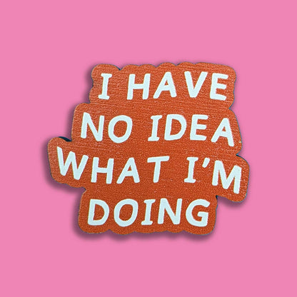 A die-cut enamel pin with the text 'I HAVE NO IDEA WHAT I'M DOING' in white lettering on a textured orange-brown background, set against a solid pink backdrop.