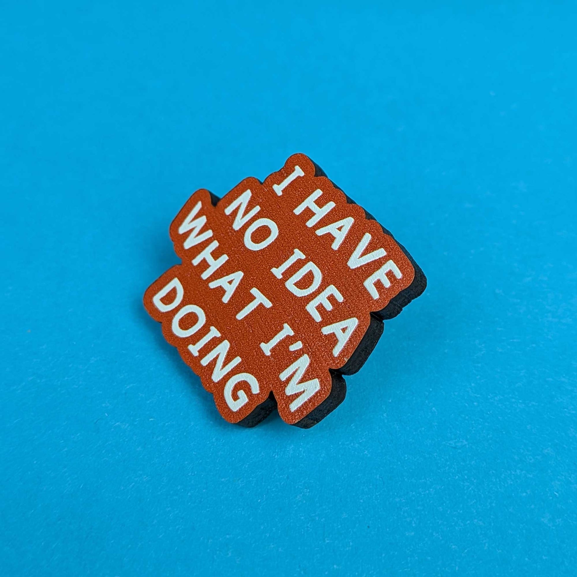 A small brown enamel pin with white text reading 'I HAVE NO IDEA WHAT I'M DOING', photographed on a bright blue background.