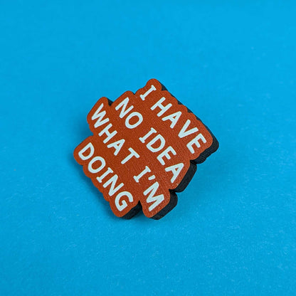 A small brown enamel pin with white text reading 'I HAVE NO IDEA WHAT I'M DOING', photographed on a bright blue background.