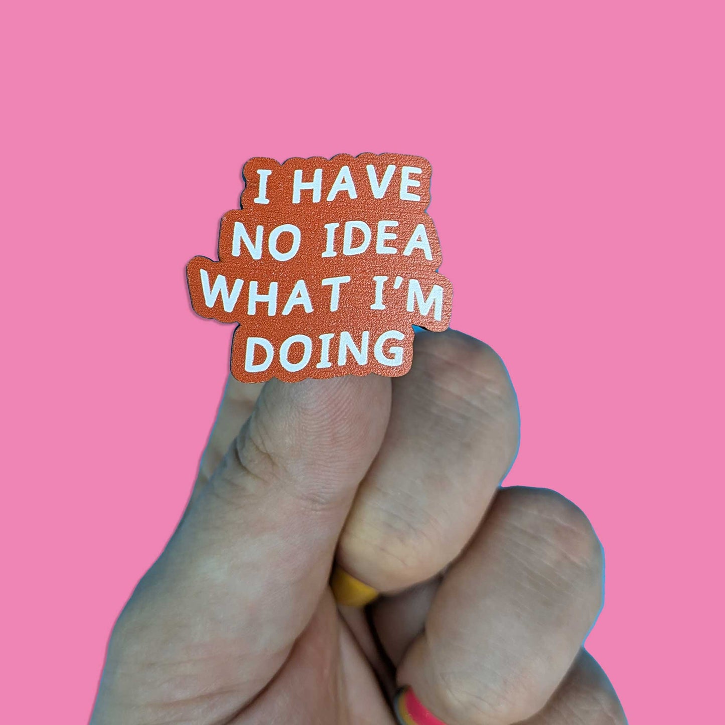 A hand holding a small die-cut sticker that reads 'I HAVE NO IDEA WHAT I'M DOING' in white text on a terracotta orange background, set against a solid pink backdrop.