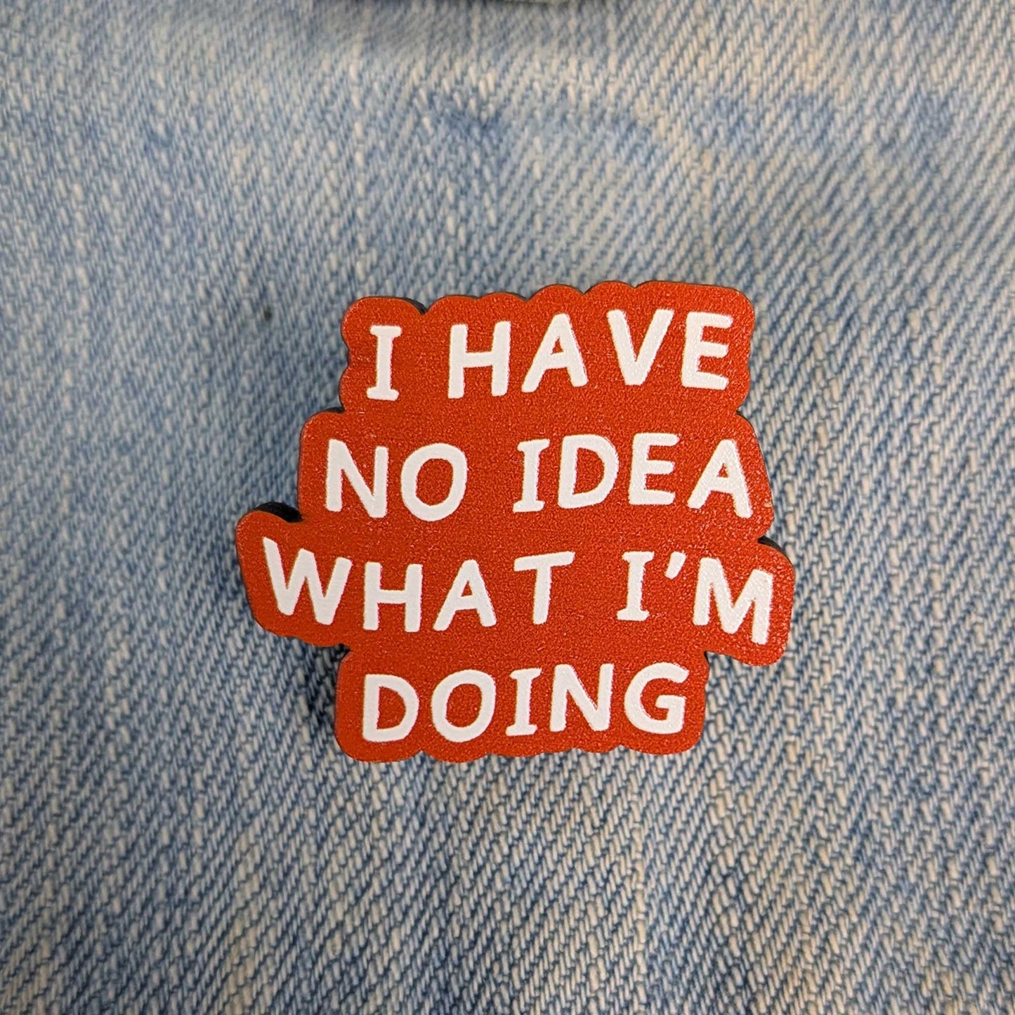 An orange-red enamel pin with white text reading 'I HAVE NO IDEA WHAT I'M DOING' placed on a light blue denim fabric background.