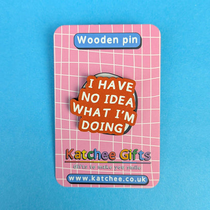 A wooden pin displayed on its pink backing card. The pin features a brown wooden base with layered orange, red, and yellow speech-bubble shapes containing the text 'I HAVE NO IDEA WHAT I'M DOING' in white.