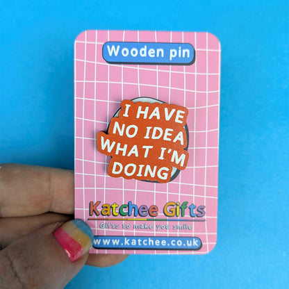 A hand holds a pink card backing displaying a wooden pin. The pin has an orange-brown wood-grain background with the text 'I HAVE NO IDEA WHAT I'M DOING' in white.