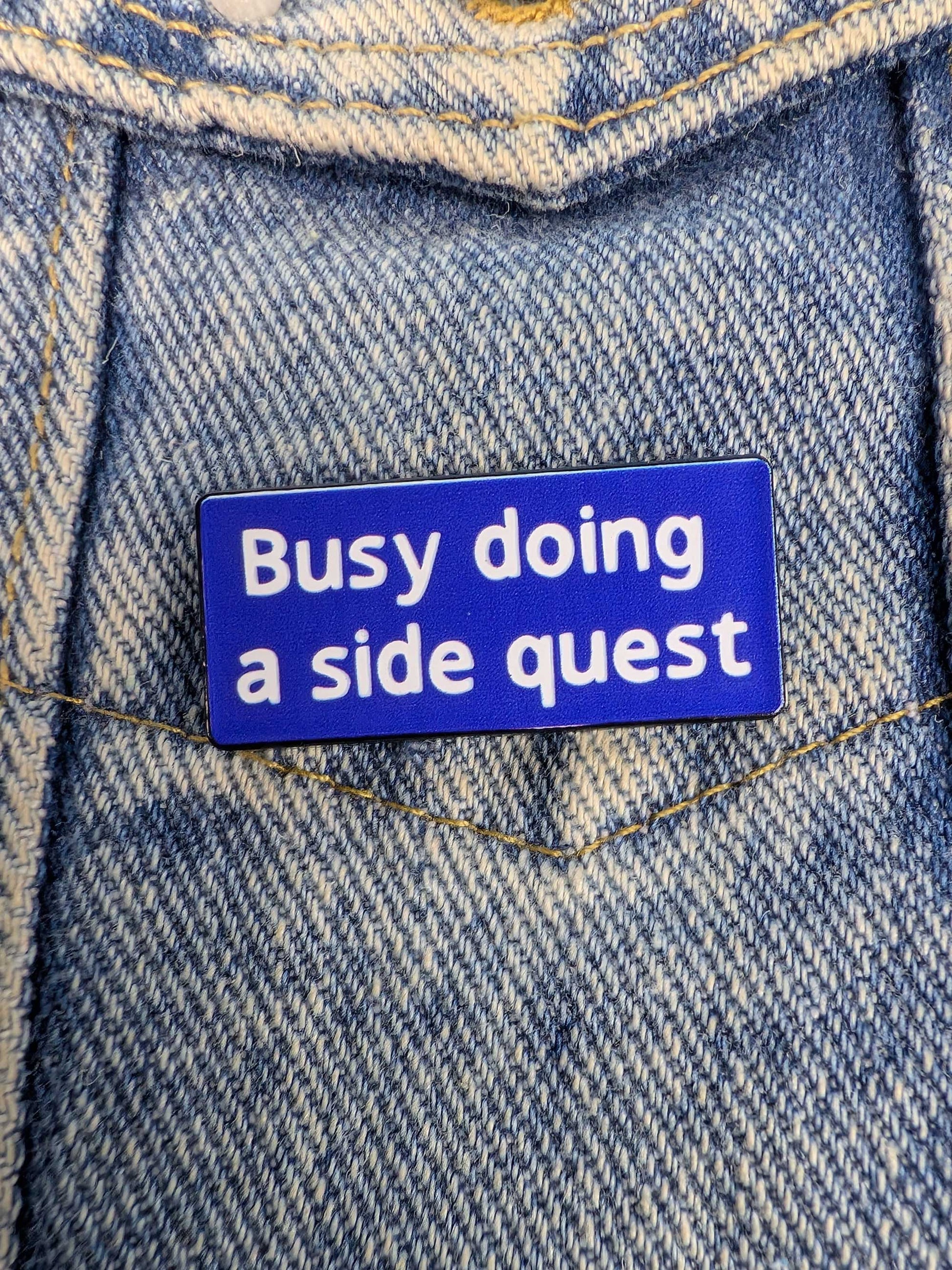 Blue enamel pin with 'Busy doing a side quest' text on a denim background