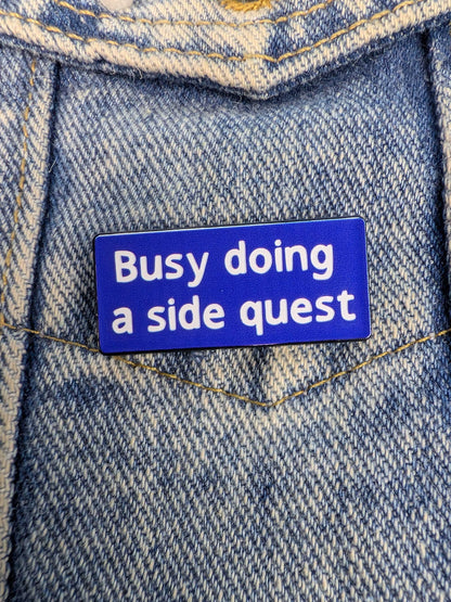 Blue enamel pin with 'Busy doing a side quest' text on a denim background