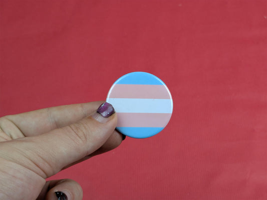 Transgender pride flag button held by a hand against a red background