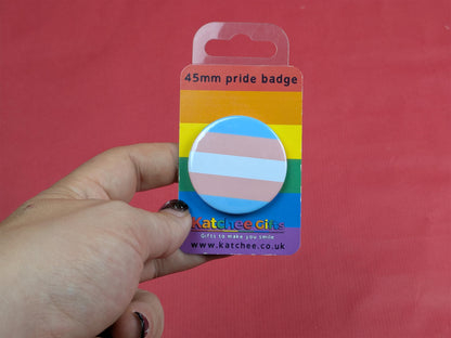 Trans pride badge in a rainbow packaging held against a red background