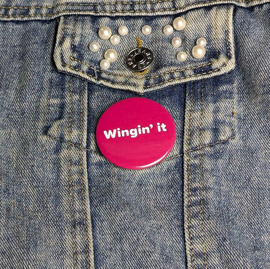 Pink button with 'Wingin' it' text on a denim jacket.